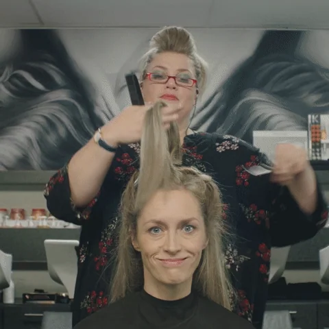 Hairdresser holding up a long section of a client's hair, scissors ready, while the client smiles nervously