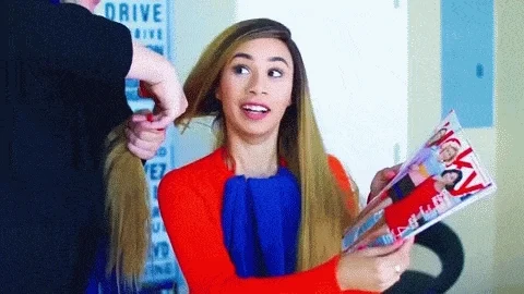 A woman holding a fashion magazine looks shocked as a hairdresser cuts off a large section of her hair.