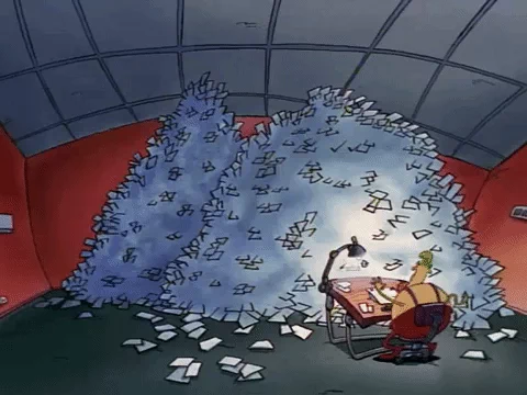 Rajzfilmfigura dolgozik az íróasztalánál egy plafonig érő papírhalom alatt.