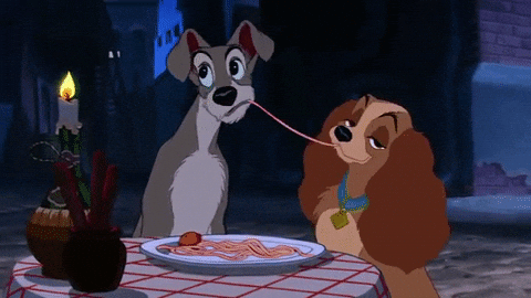 A scene from the Disney movie, Lady and the Tramp. Lady and the Tramp are seated at a small table, facing away from each other. They each take bites of the same long strand of spaghetti. As they chew, they inadvertently lean in, and their lips meet in a kiss